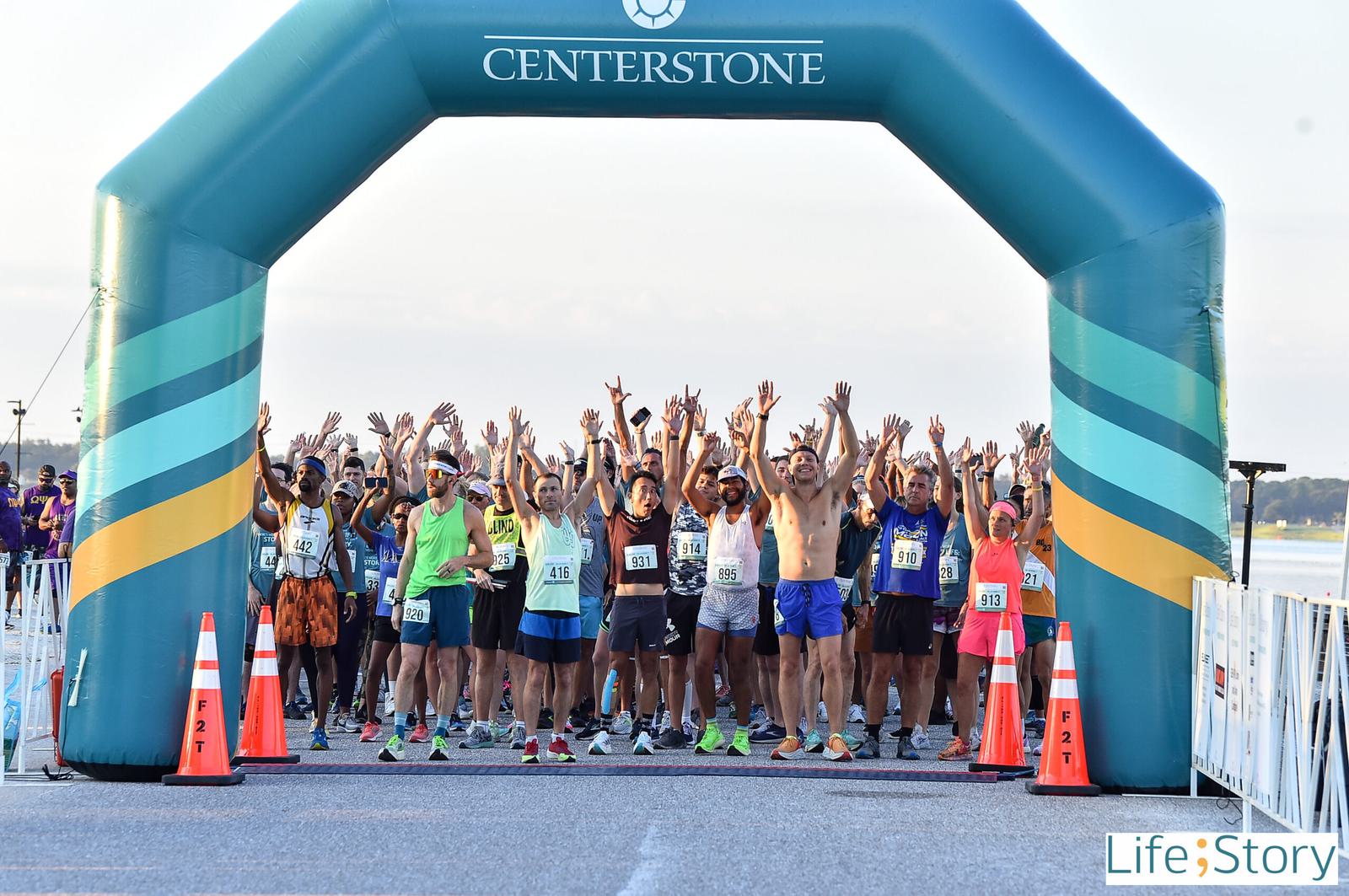 A large group of runners stands behind a blue inflatable arch with "Centerstone" on it, raising their arms in the air at the start of a race. Orange cones and spectators line the sides.