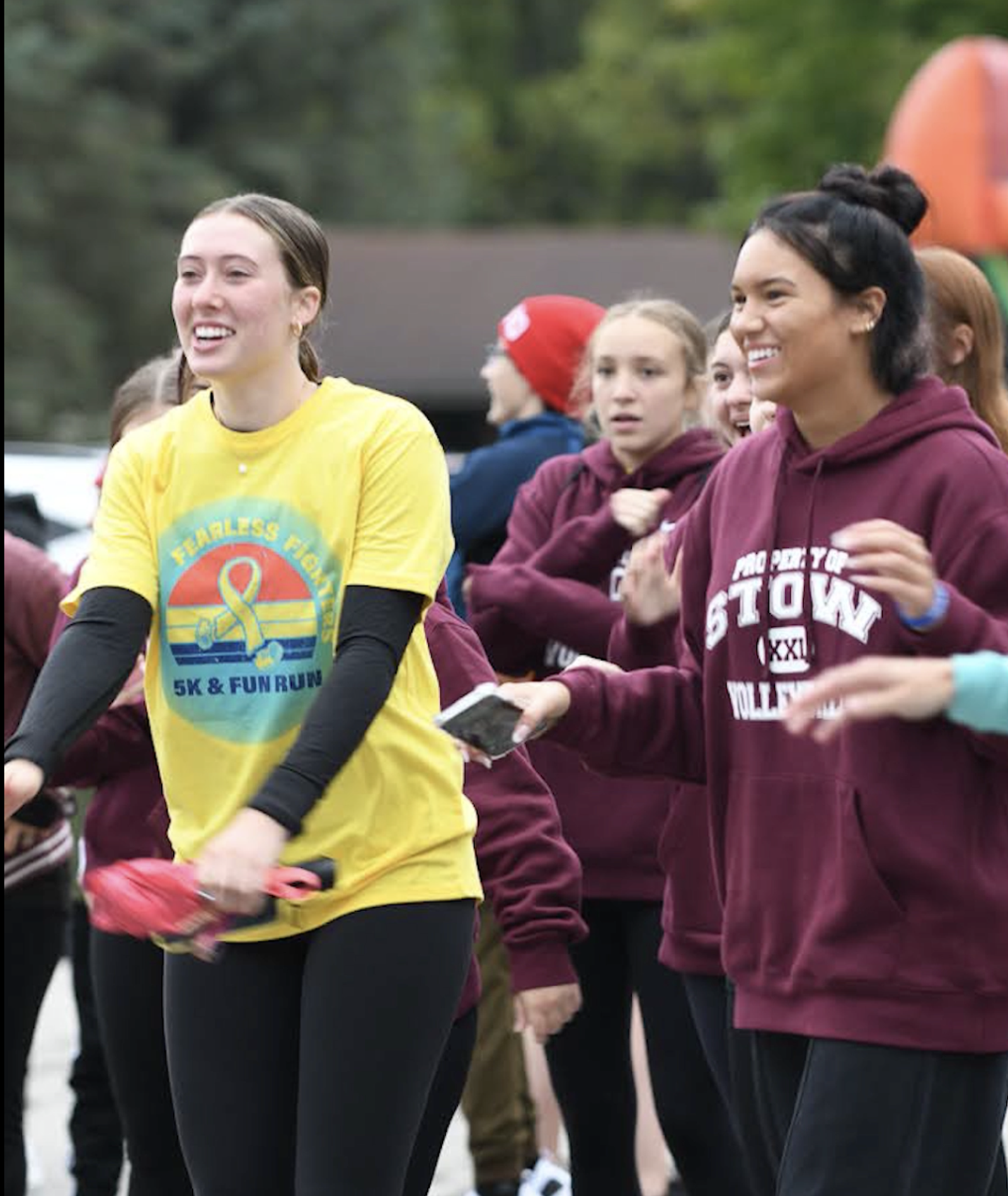 Two young women, one in a yellow "5K & Fun Run" shirt and the other in a maroon hoodie, smile and walk with a group of people outdoors, appearing happy and energetic during an event.