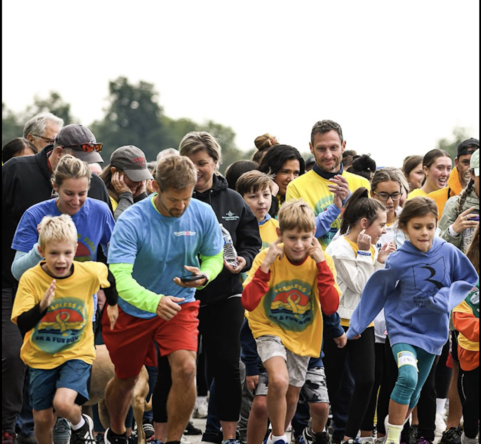 A group of adults and children, some wearing race bibs and colorful shirts, eagerly start running at an outdoor fun run event, with trees visible in the background.
