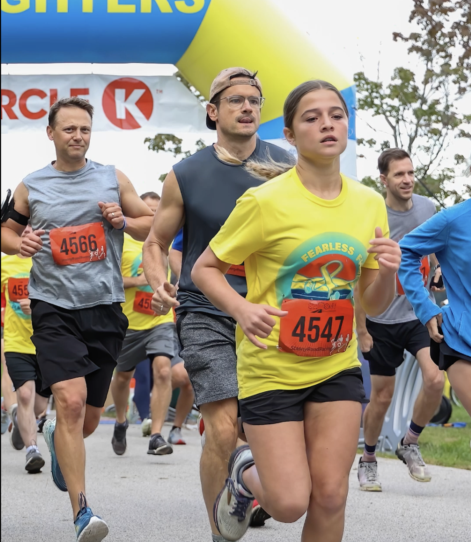 A group of runners, including a girl in a yellow "Fearless" shirt (bib 4547), are participating in a race. Other runners wear athletic gear and numbered bibs. A blue and yellow inflatable arch is visible in the background.