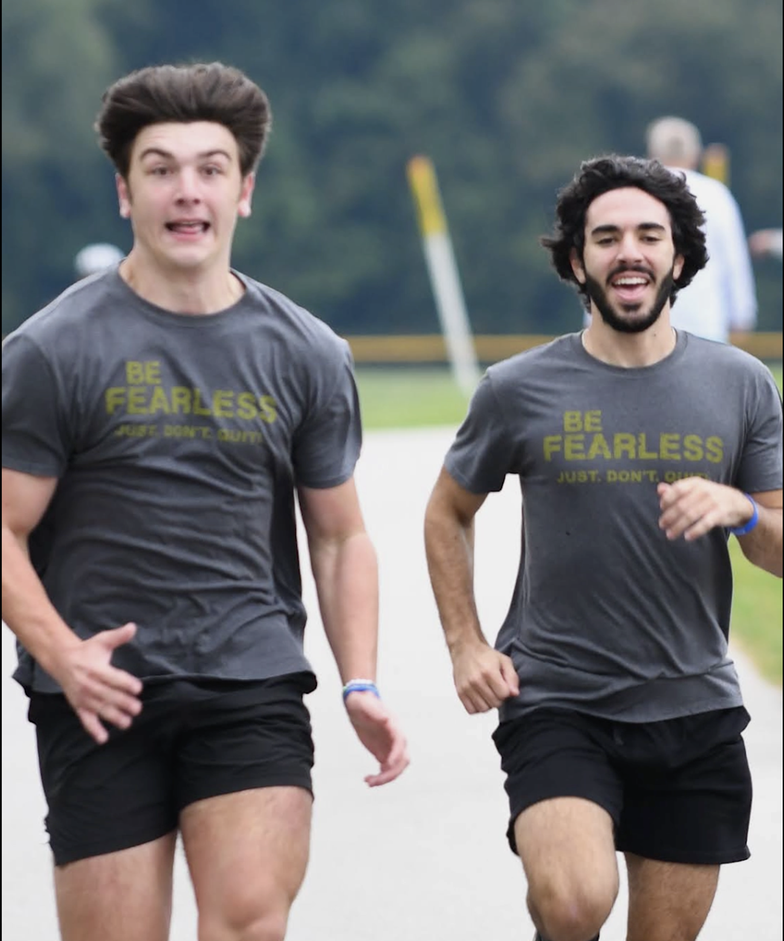 Two young men in matching "Be Fearless" shirts and black shorts are running outdoors on a paved path, both smiling and enjoying the run, with a blurry background of grass and trees.