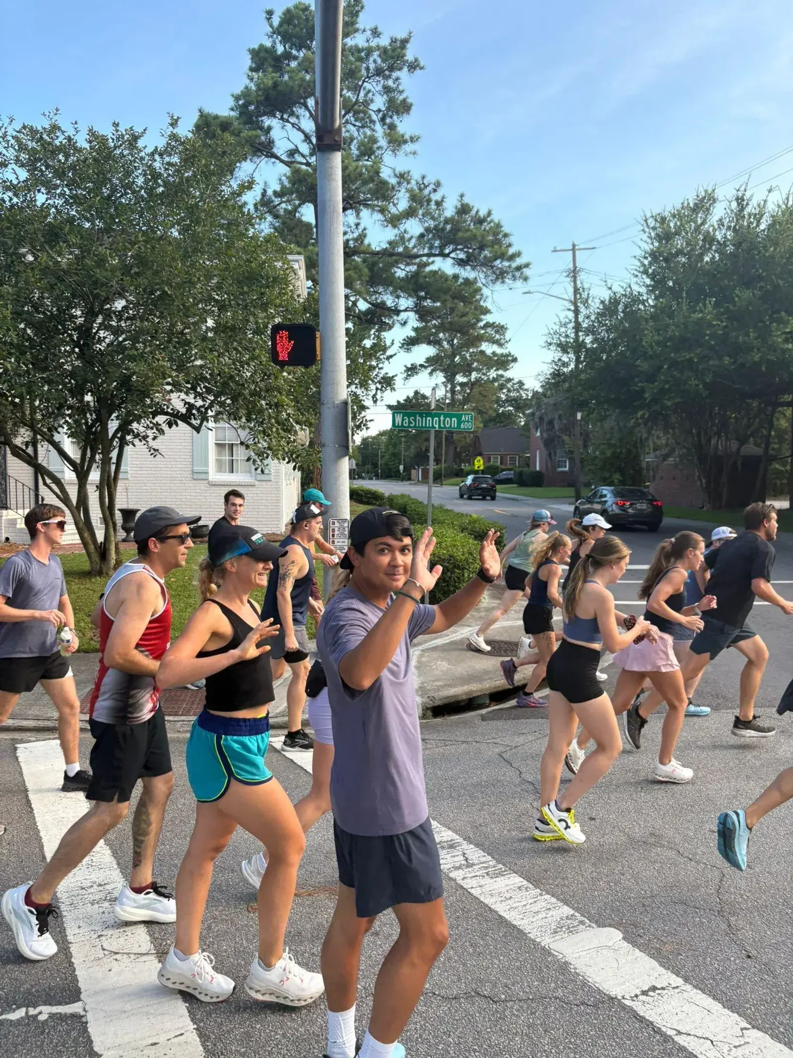 A group of people jog together through a crosswalk on a sunny day. Most are wearing athletic clothes, and some wave at the camera. Trees, houses, and a street sign for Washington Ave are visible in the background.