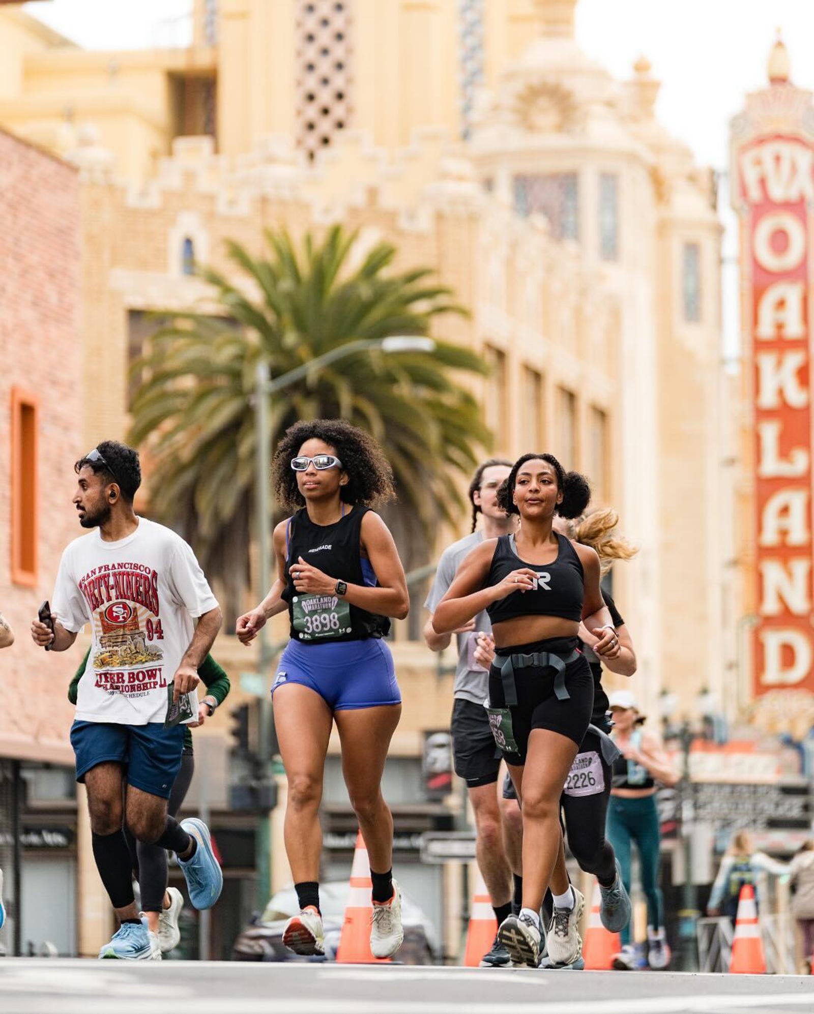 A group of runners participate in a race through a city street with palm trees and a vintage “Oakland” theater sign visible in the background. The runners look focused and energetic.