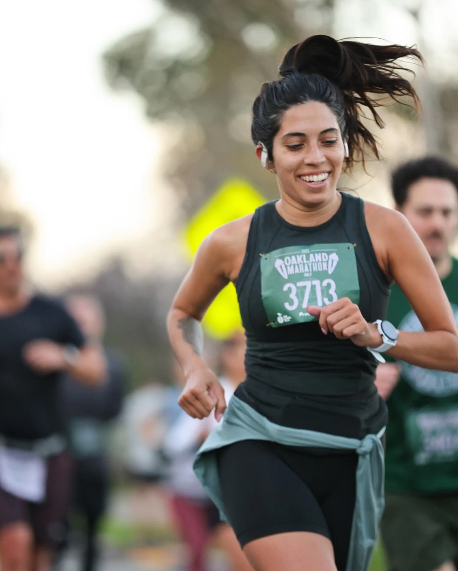 A woman smiles while running in a marathon, wearing a bib labeled "Oakland Marathon 3713." Other runners are visible in the blurred background outdoors.
