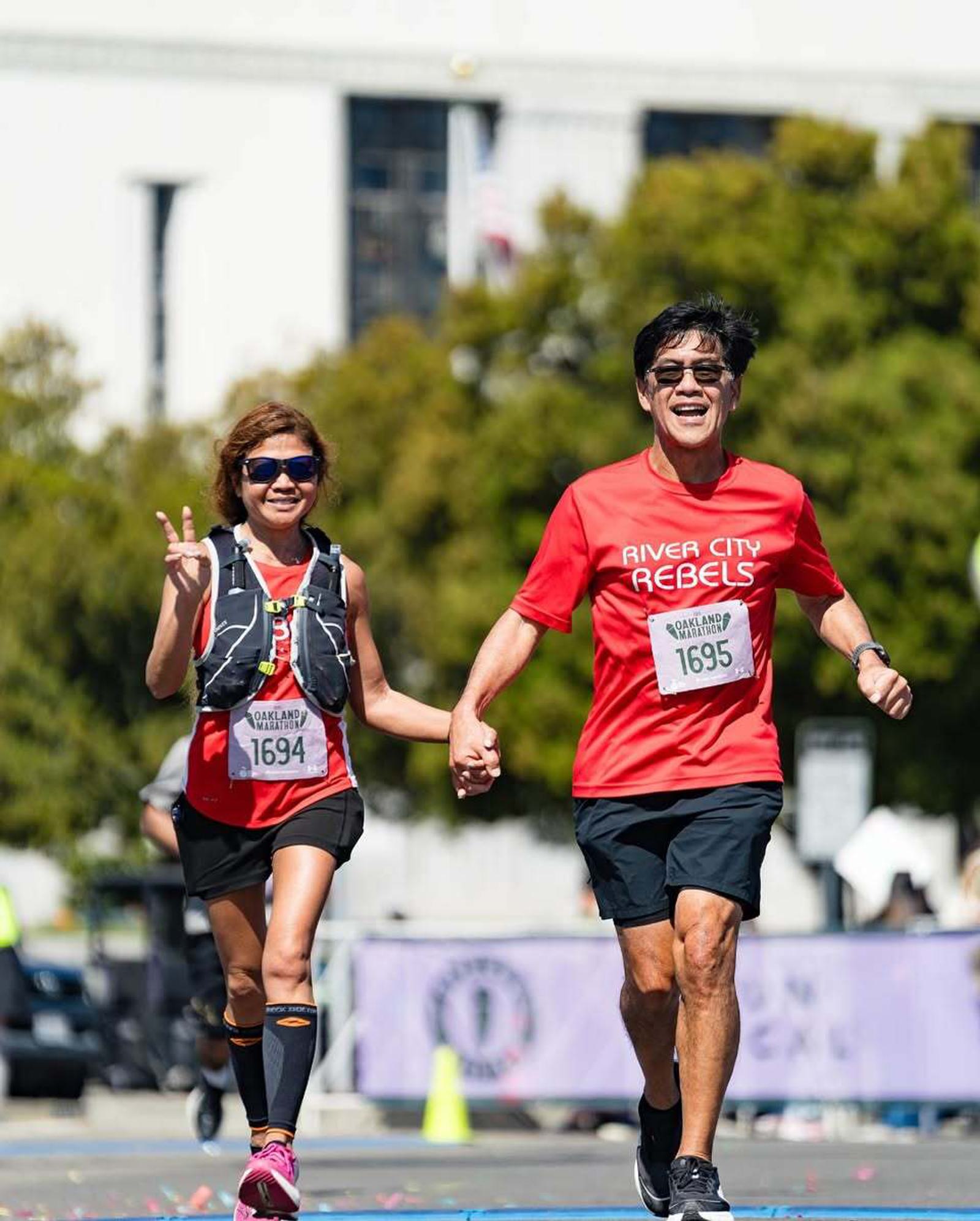 Two smiling runners wearing red “River City Rebels” shirts hold hands while crossing a race finish line outdoors. The woman gives a peace sign, and both wear sunglasses and race bibs numbered 1694 and 1695.