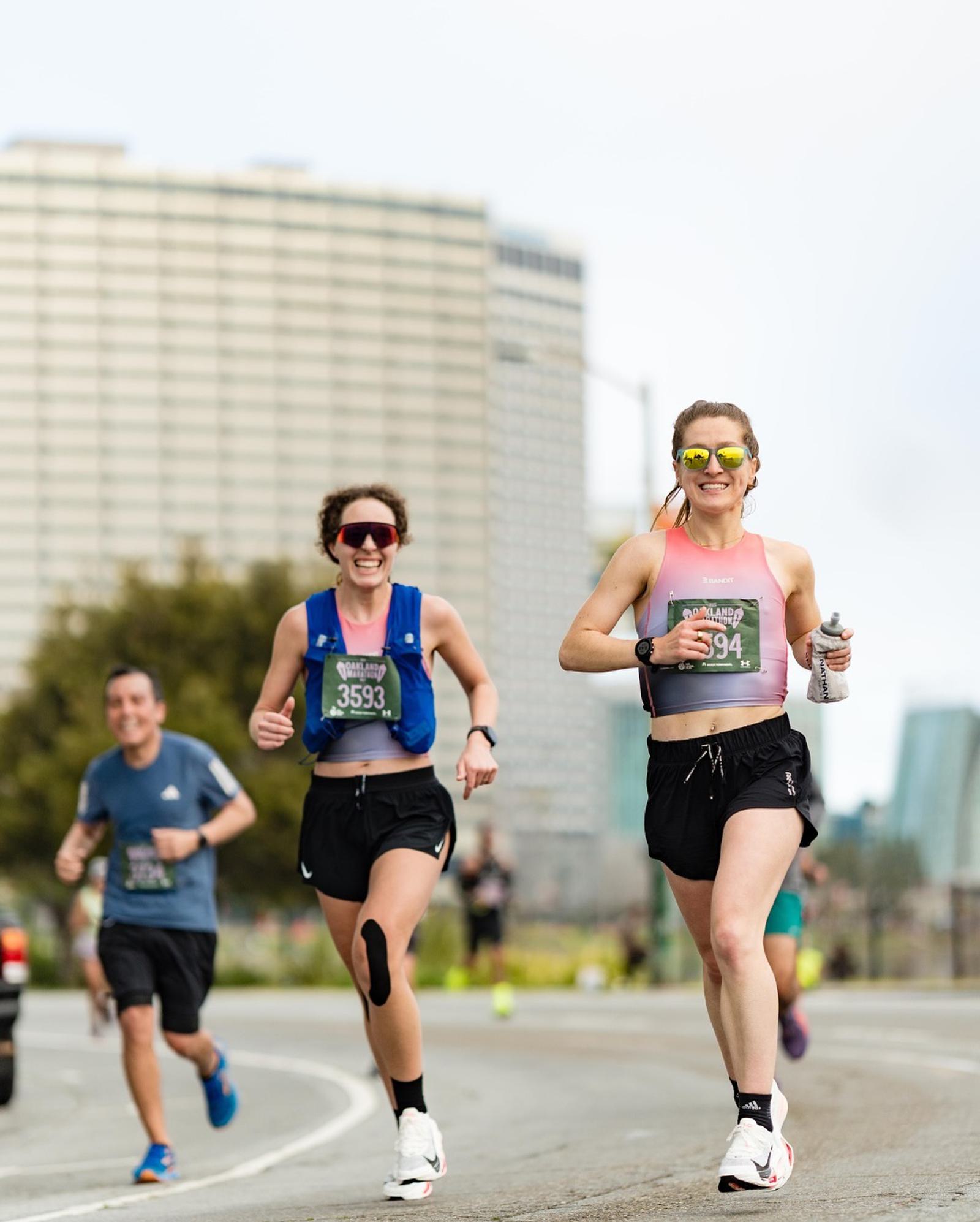 Two women in athletic gear smile and run in a road race, each wearing race bibs and sunglasses. A man and another participant run behind them on a city street with tall buildings in the background.