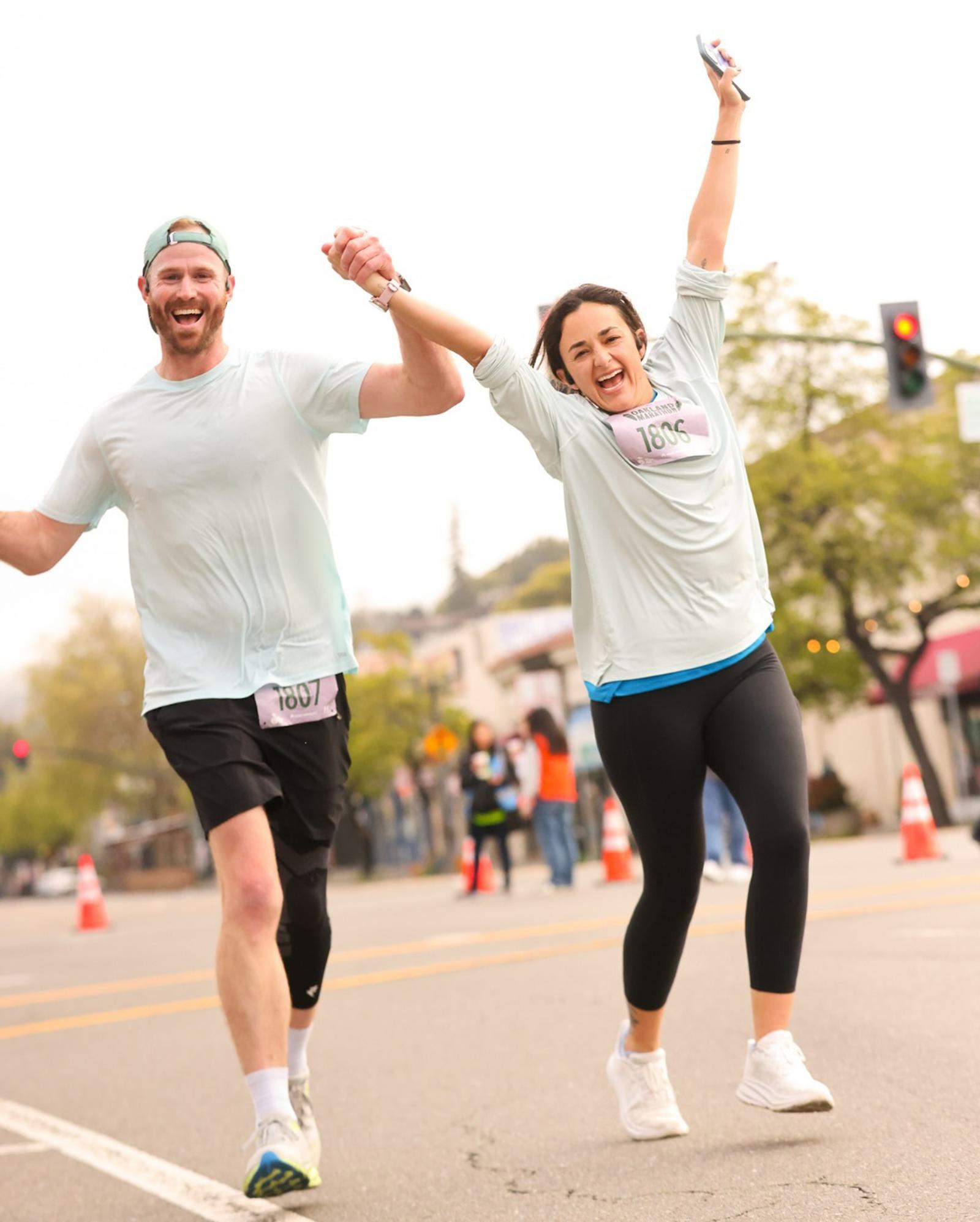 Two runners, a man and a woman, hold hands and celebrate as they cross a city street during a race. Both are smiling and wearing light shirts with race bibs. The woman raises one arm joyfully, and there are cones and people in the background.