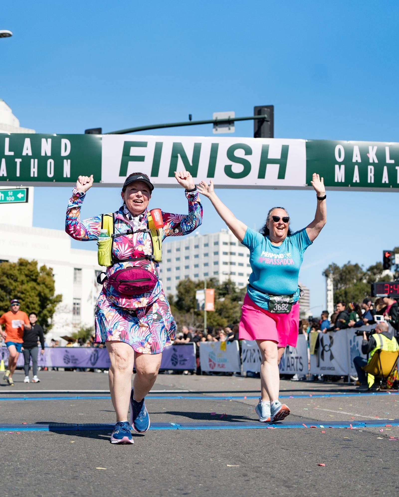 Two women joyfully raise their arms as they cross the finish line at a marathon event. Both are smiling and wearing colorful athletic clothing, with the FINISH sign and cheering spectators in the background.