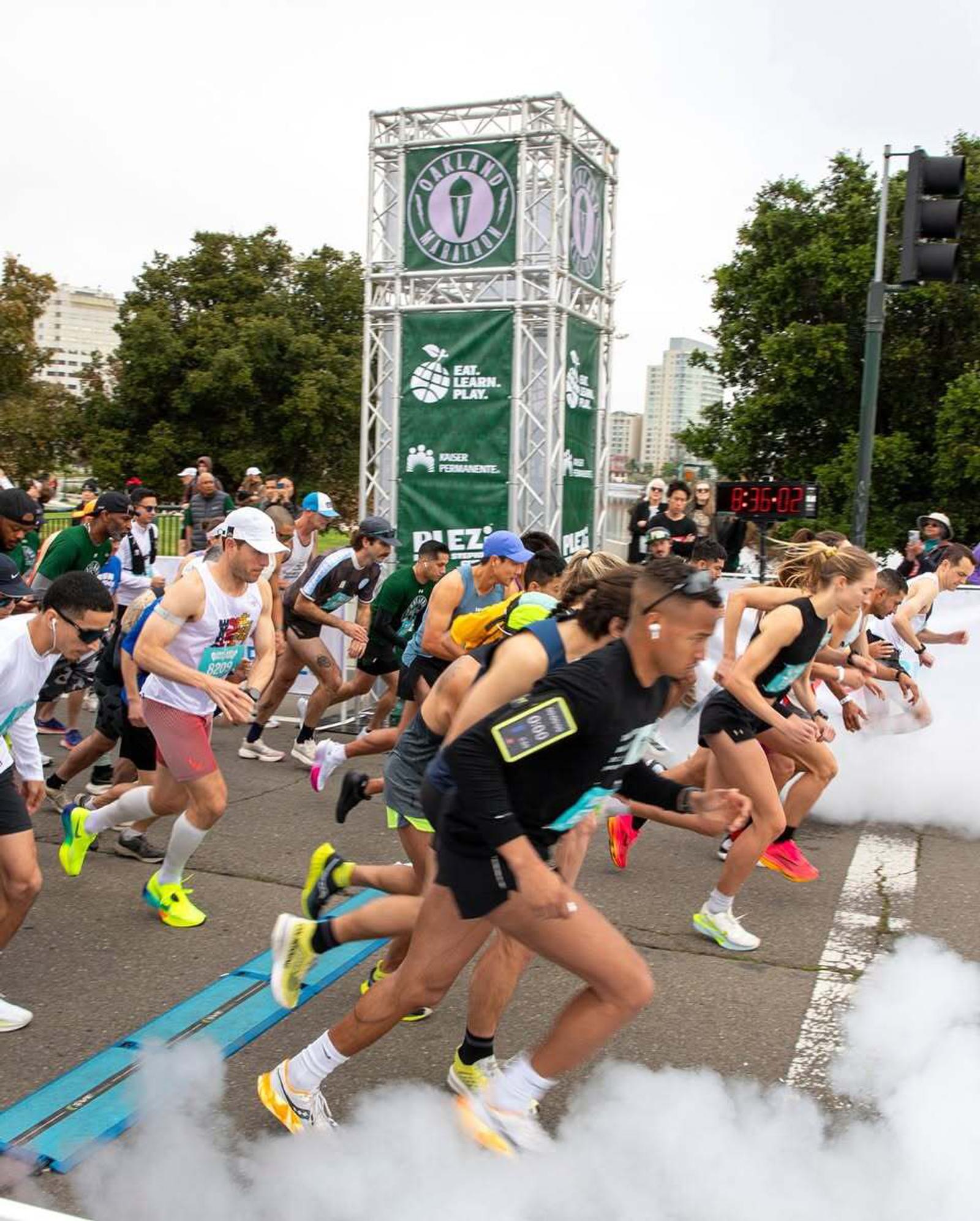 A group of runners in athletic gear start a race on a city street, with a large event tower, digital timer, and spectators in the background. White smoke rises at the starting line. Trees and buildings are visible behind them.