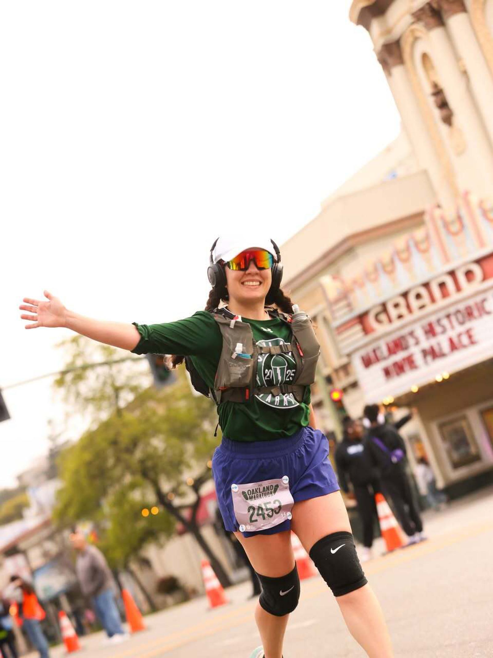A runner wearing sunglasses, headphones, and a dark green shirt with a race bib smiles and spreads her arms wide as she runs in a city street race. A theater marquee and spectators are visible in the background.