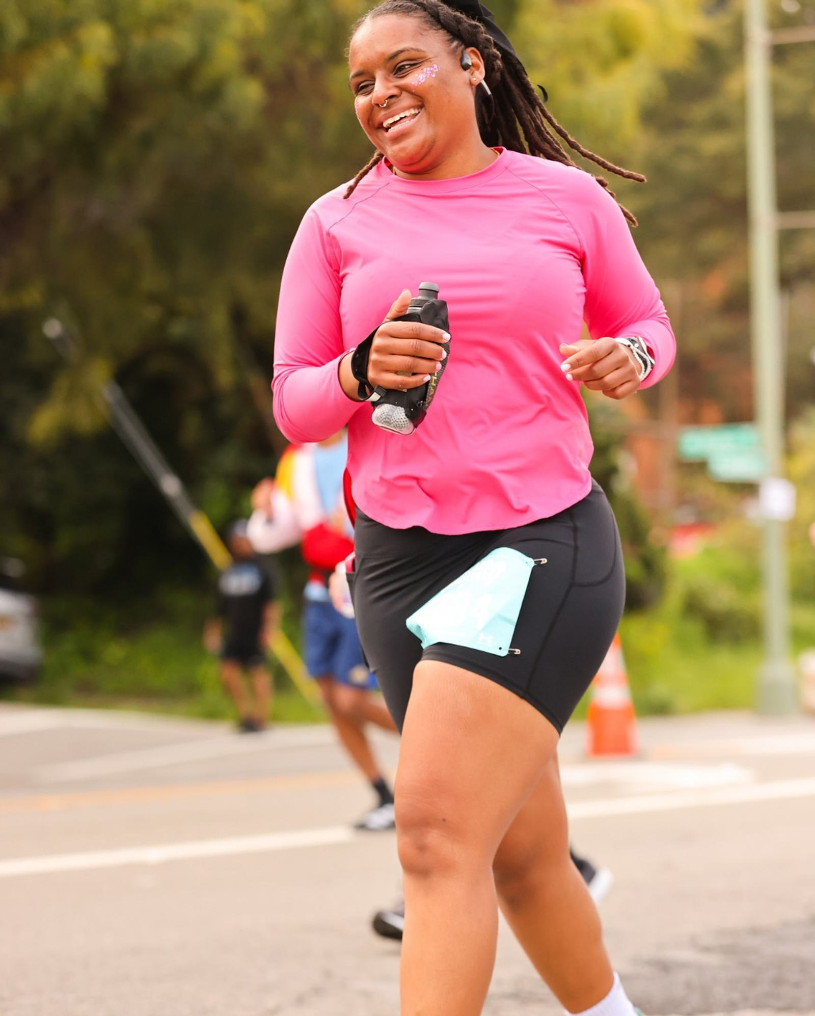 A woman smiling and running outdoors in athletic wear, holding a water bottle and wearing a pink long-sleeve shirt and black shorts with a blue cloth tucked in. Trees and other runners are visible in the background.