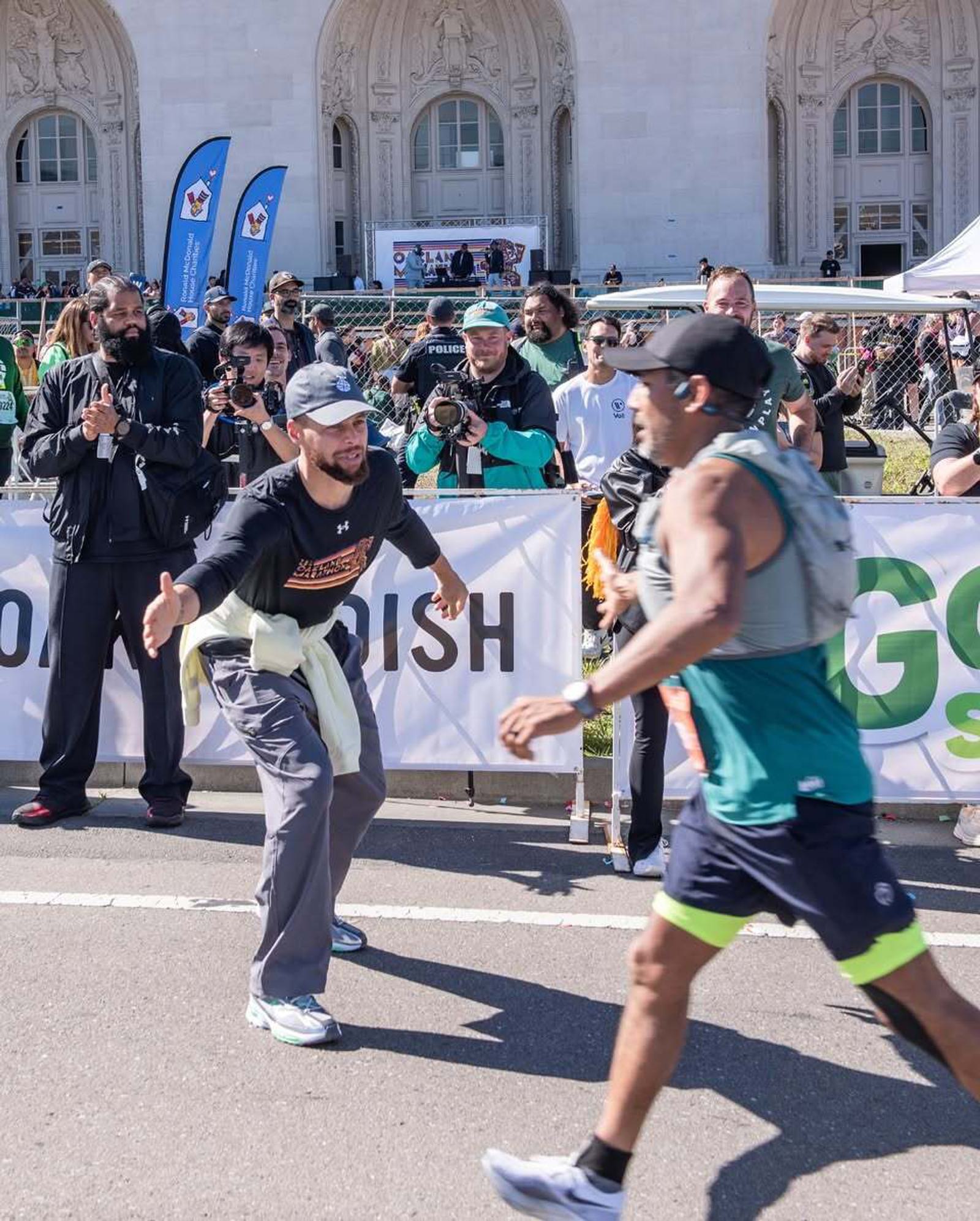 A man in athletic wear reaches out to congratulate a marathon runner near the finish line, with a cheering crowd and photographers behind a barrier in front of an ornate building.