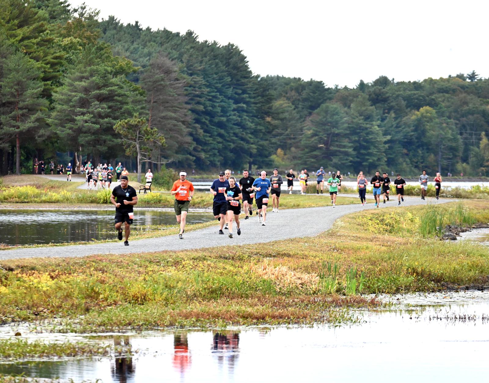 A group of people are running a race on a gravel path beside a pond, surrounded by green trees and grass on a cloudy day.