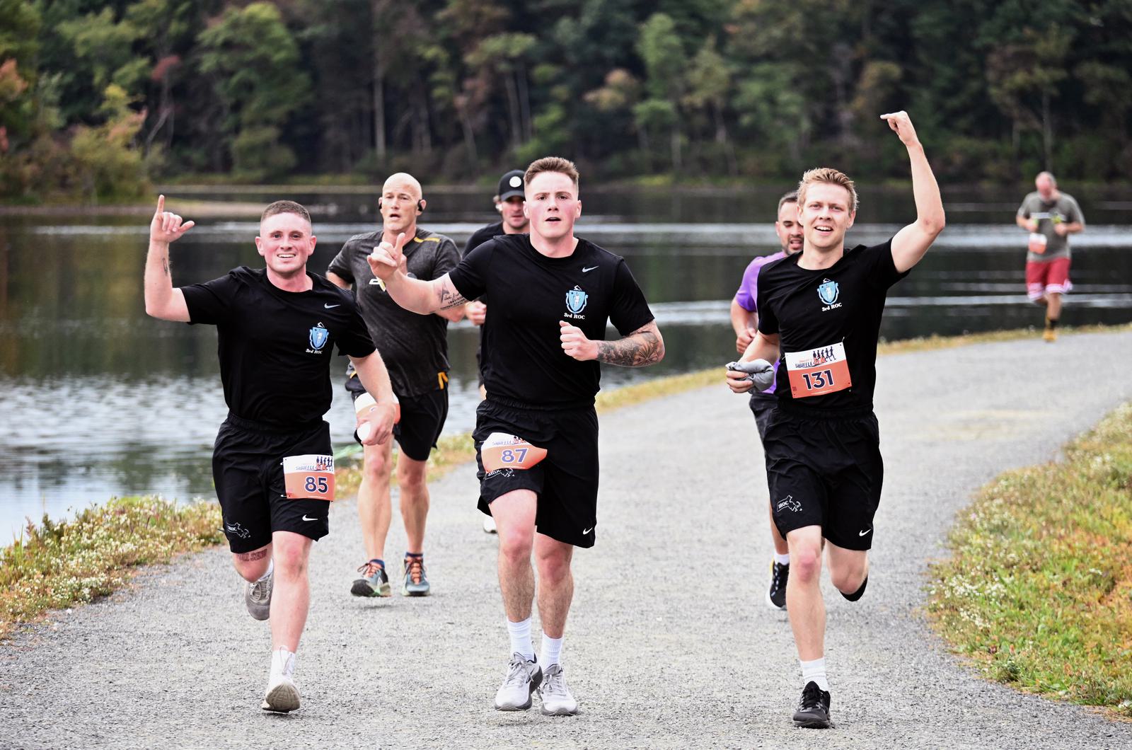 Three men in black shirts and race bibs run and cheer on a lakeside path, while other runners follow behind. Trees and water are in the background, and everyone appears energetic and happy.