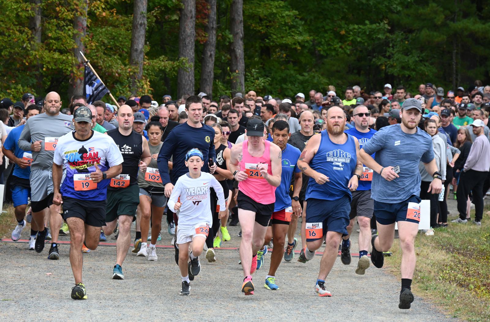 A large group of people of various ages start a running race on a gravel path in a wooded area, wearing numbered bibs and athletic gear, with trees and more runners in the background.