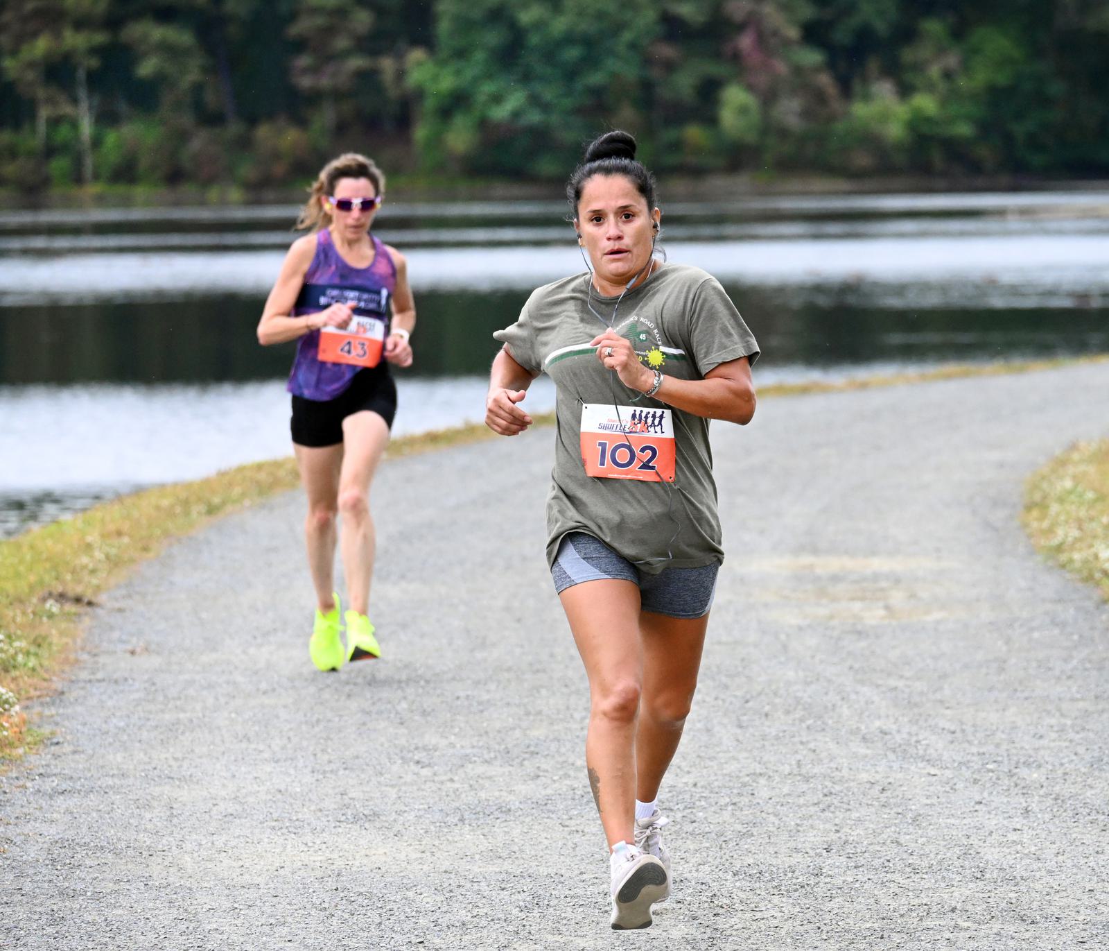 Two women are running on a gravel path next to a lake during a race, both wearing race bibs and athletic clothes. The woman in front, wearing bib 102, looks determined, while the second runner follows close behind.