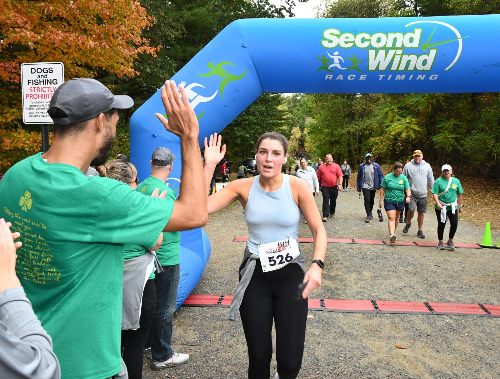 A woman wearing running bib 526 high-fives a man in a green shirt as she finishes a race under a blue “Second Wind Race Timing” arch. Several people walk behind her on the wooded trail.