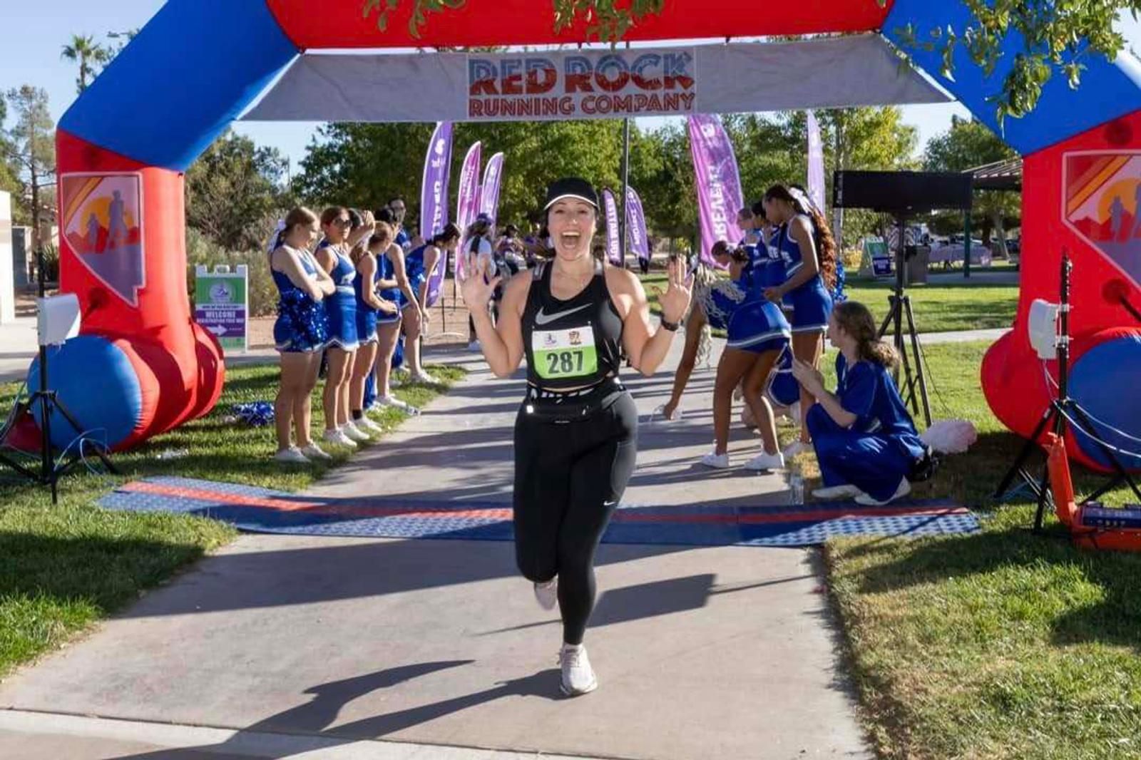 A runner wearing bib number 287 smiles and raises both hands in celebration as she crosses the finish line at a Red Rock Running Company event, with cheerleaders applauding on both sides under a blue and red inflatable arch.