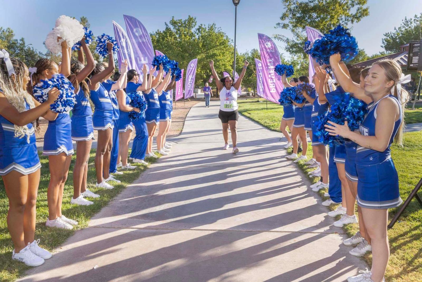 A runner raises her arms in victory as she crosses a finish line, cheered on by two rows of blue-uniformed cheerleaders holding pom-poms along a sunny path lined with purple flags.