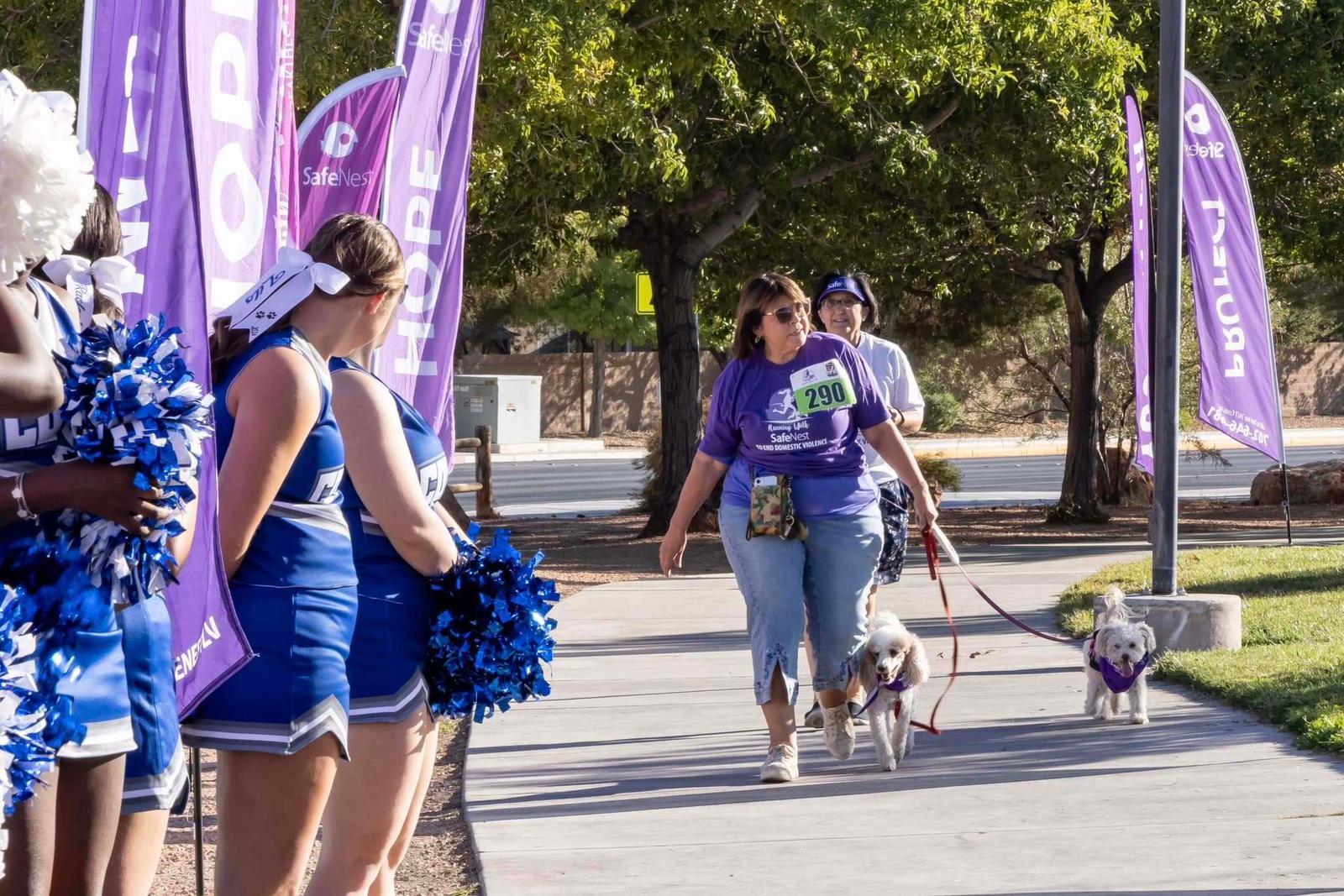 A woman wearing a purple shirt and numbered race bib walks two small dogs on leashes along a path lined with purple "HOPE" banners, while cheerleaders in blue uniforms watch and cheer from the side.