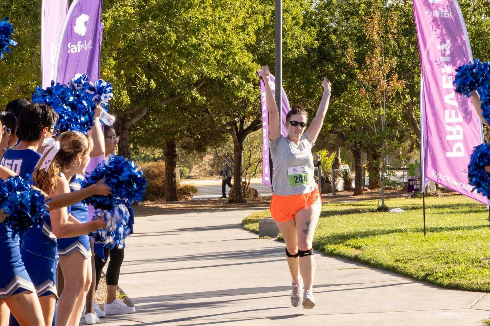 A runner in orange shorts and a gray shirt crosses the finish line of a race with arms raised in victory, while cheerleaders with blue pom-poms cheer on both sides. Pink banners and trees are visible in the background.