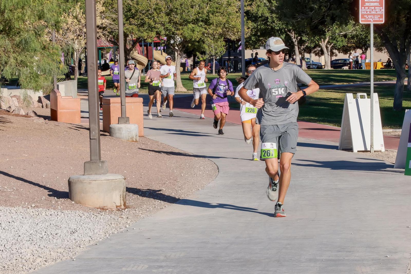 A group of runners participate in a 5K race on a paved path in a park, with one runner in a gray outfit leading and others following behind under sunny, clear skies. Trees and park signs are visible in the background.