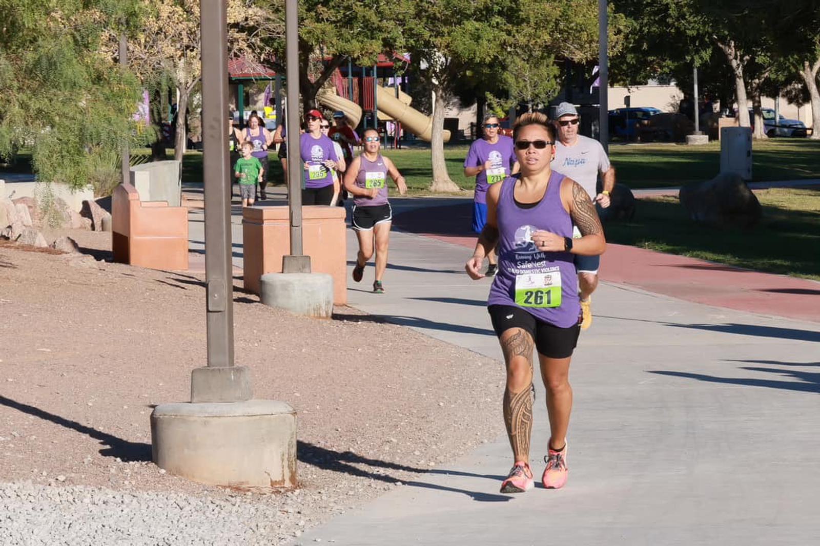 A group of people run outdoors on a paved park path during a race. The leading runner, wearing a purple tank top and black shorts, has a tattooed leg and wears bib number 261. Trees and playground equipment are in the background.