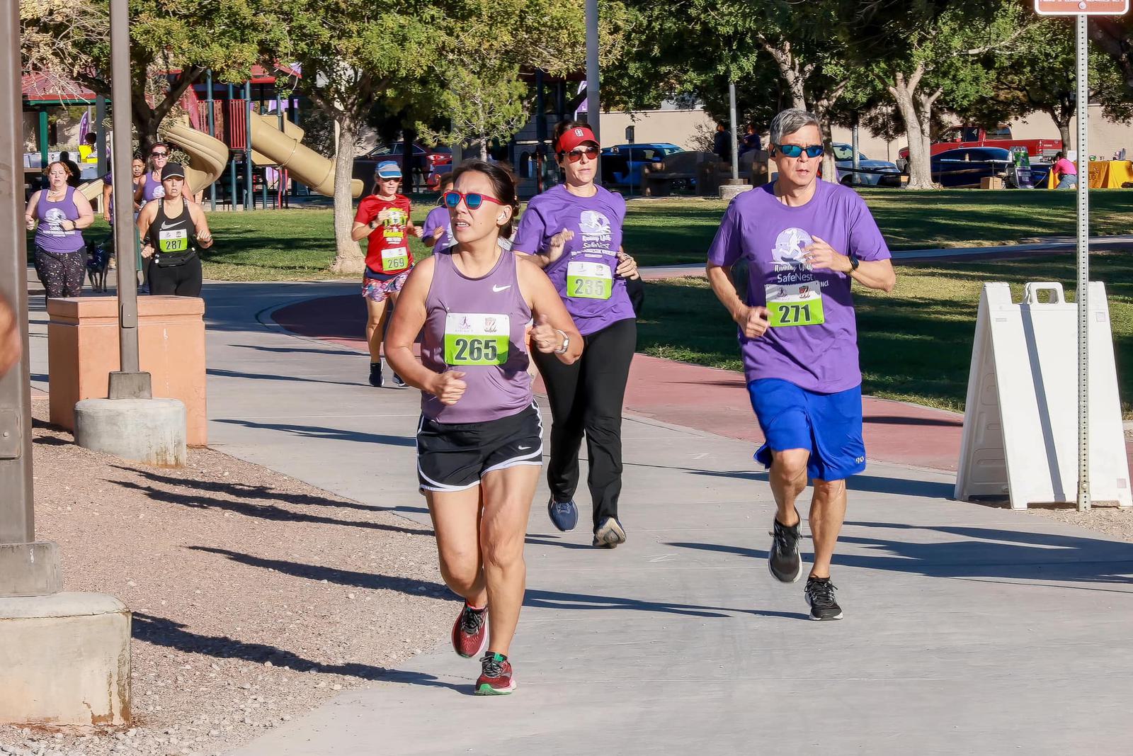 Runners wearing numbered bibs and purple shirts participate in a race at a park on a sunny day, with trees and playground equipment visible in the background.