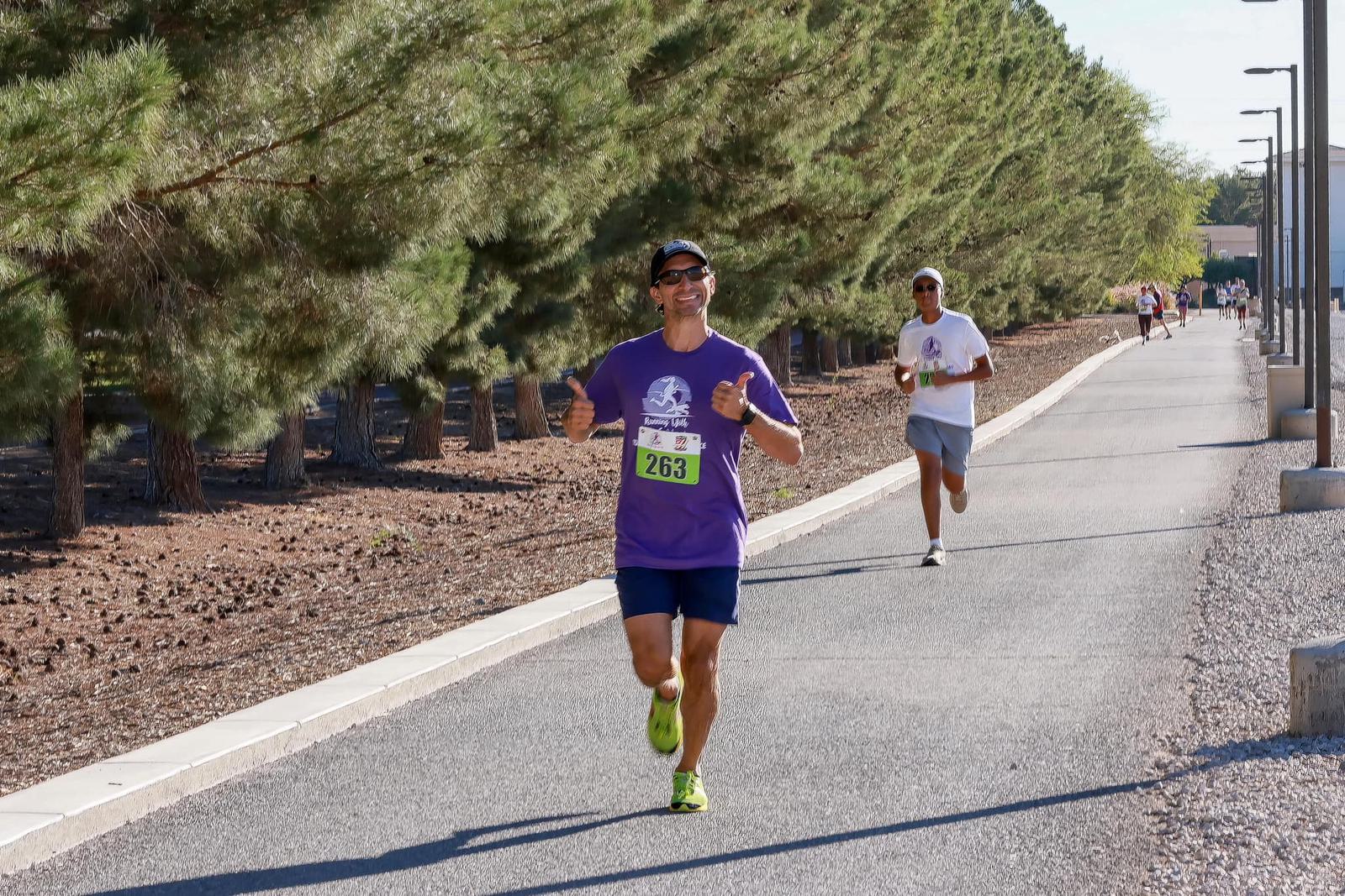 A man in a purple shirt and sunglasses runs and smiles, giving a thumbs-up on a paved path lined with trees, while another runner follows behind. Both are participating in a race, wearing numbered bibs.