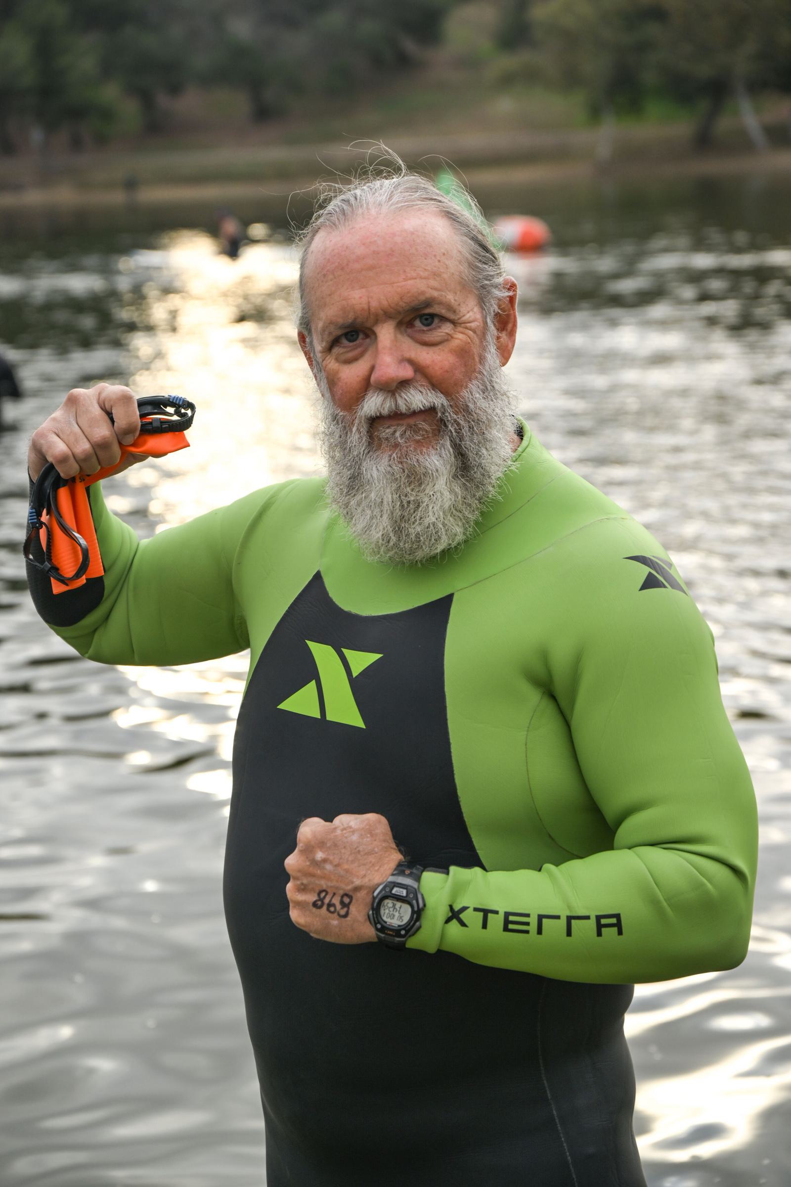 An older man with a gray beard stands in water wearing a green and black XTERRA wetsuit. He holds orange swim goggles and shows a watch on his wrist, with "858" marked on his hand. Trees and swimmers are visible in the background.