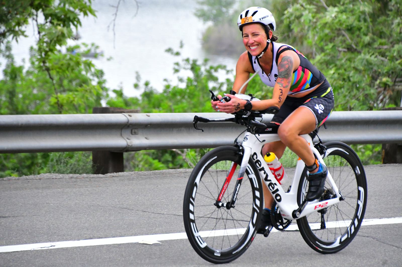 A smiling cyclist wearing a helmet and colorful triathlon suit rides a white Cervélo bike on a paved road with a guardrail and greenery in the background.