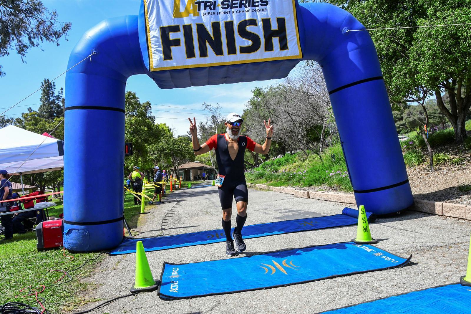 A male athlete in a black triathlon suit and sunglasses crosses the finish line of a race, raising both hands in a victory gesture under a large blue inflatable arch that reads "FINISH.