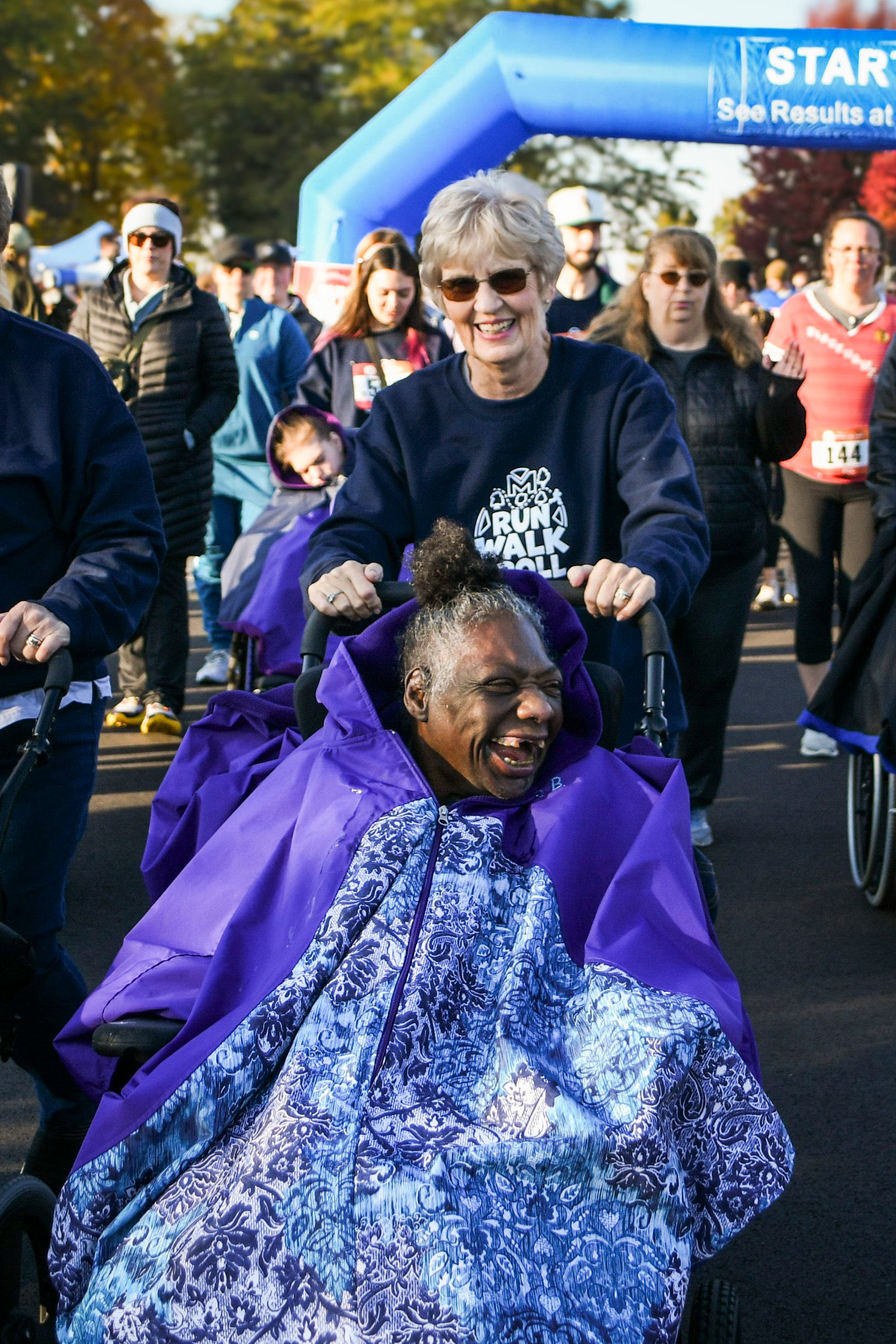 A smiling woman pushes another joyful woman in a wheelchair, both covered in purple blankets, during an outdoor group walk event on a sunny day. Several participants and a blue start/finish arch are visible in the background.