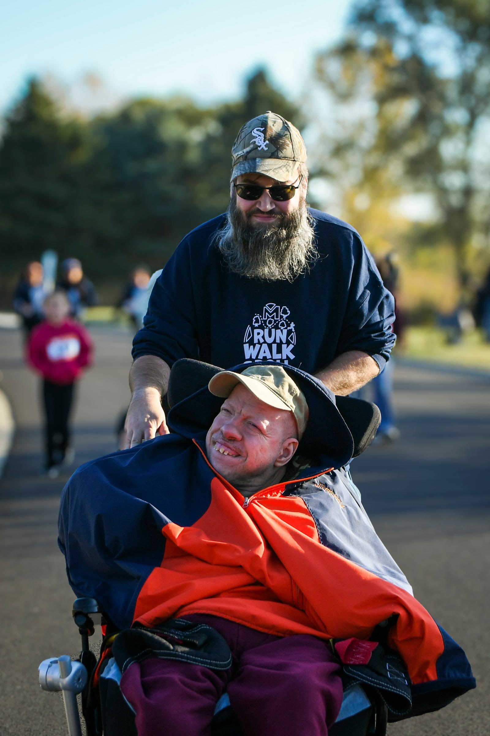 A bearded man in a cap smiles while pushing a person in a wheelchair, who is bundled in a coat and wide-brimmed hat, during an outdoor event. People and trees are visible in the background.