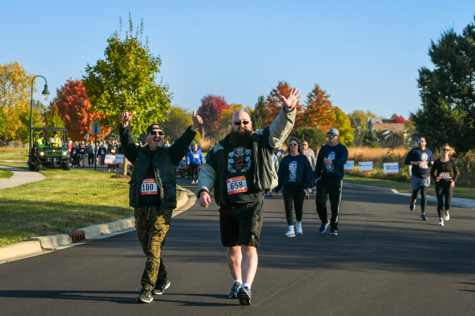 Two men with race bibs walk on a road during a race event, raising their arms and smiling. Other participants walk behind them. It's a sunny day with colorful autumn trees in the background.