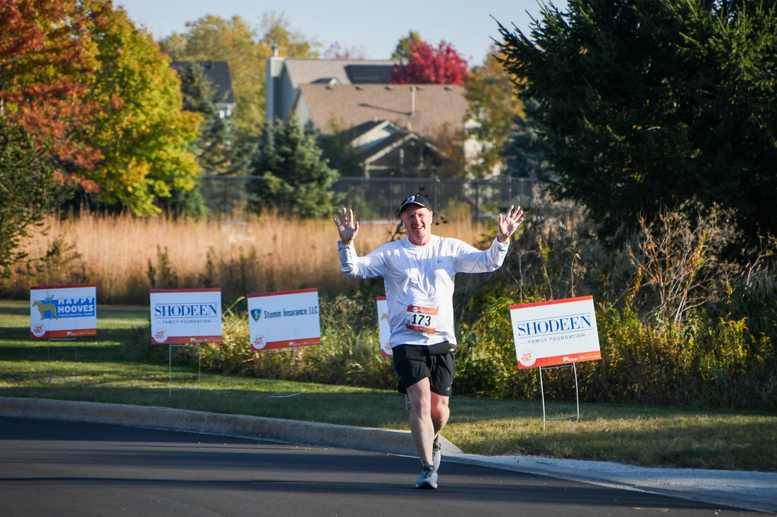 A runner wearing a white long-sleeve shirt and hat, with bib number 173, raises both arms and smiles while running on a road lined with sponsor signs and autumn trees in the background.