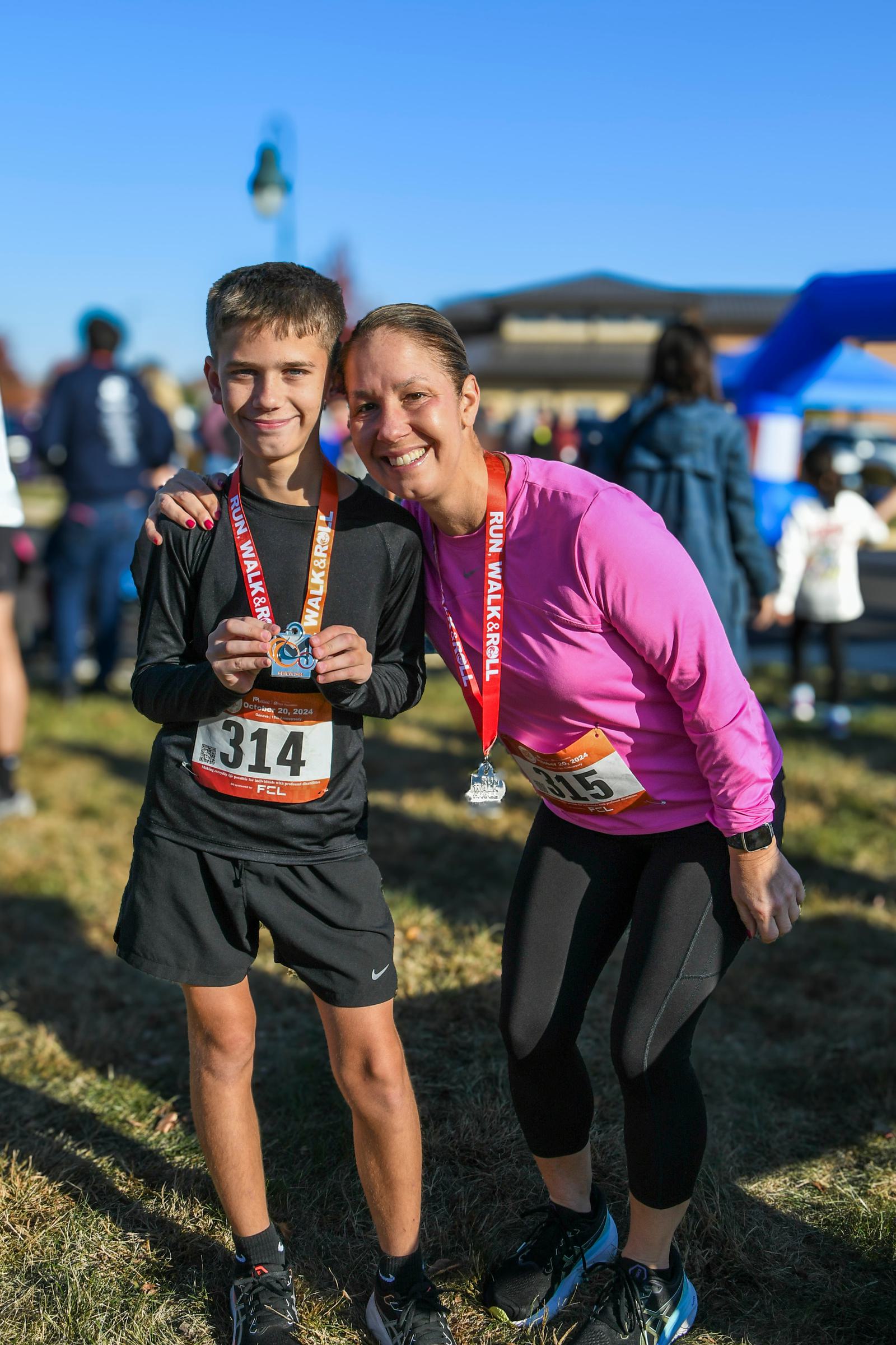 A smiling woman in a pink top and a boy in black athletic gear pose together outdoors, each wearing a race medal and bib after finishing a race event. Other participants and a blue inflatable arch are visible in the background.
