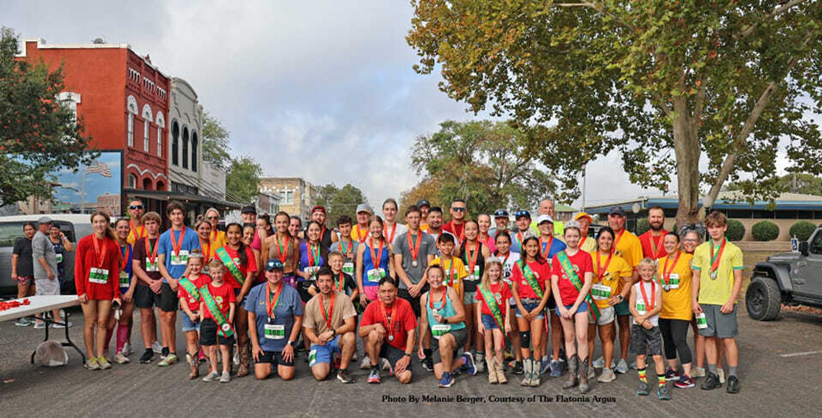 A large group of runners wearing medals and colorful shirts pose together on a city street after a race. Trees, buildings, and a jeep are visible in the background under a partly cloudy sky.