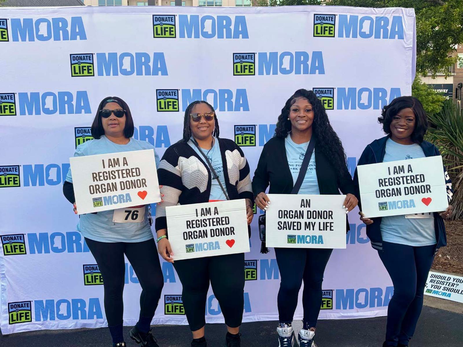 Four women stand in front of a "Donate Life MORA" backdrop, holding signs that say "I am a registered organ donor" and "An organ donor saved my life," supporting organ donation awareness.