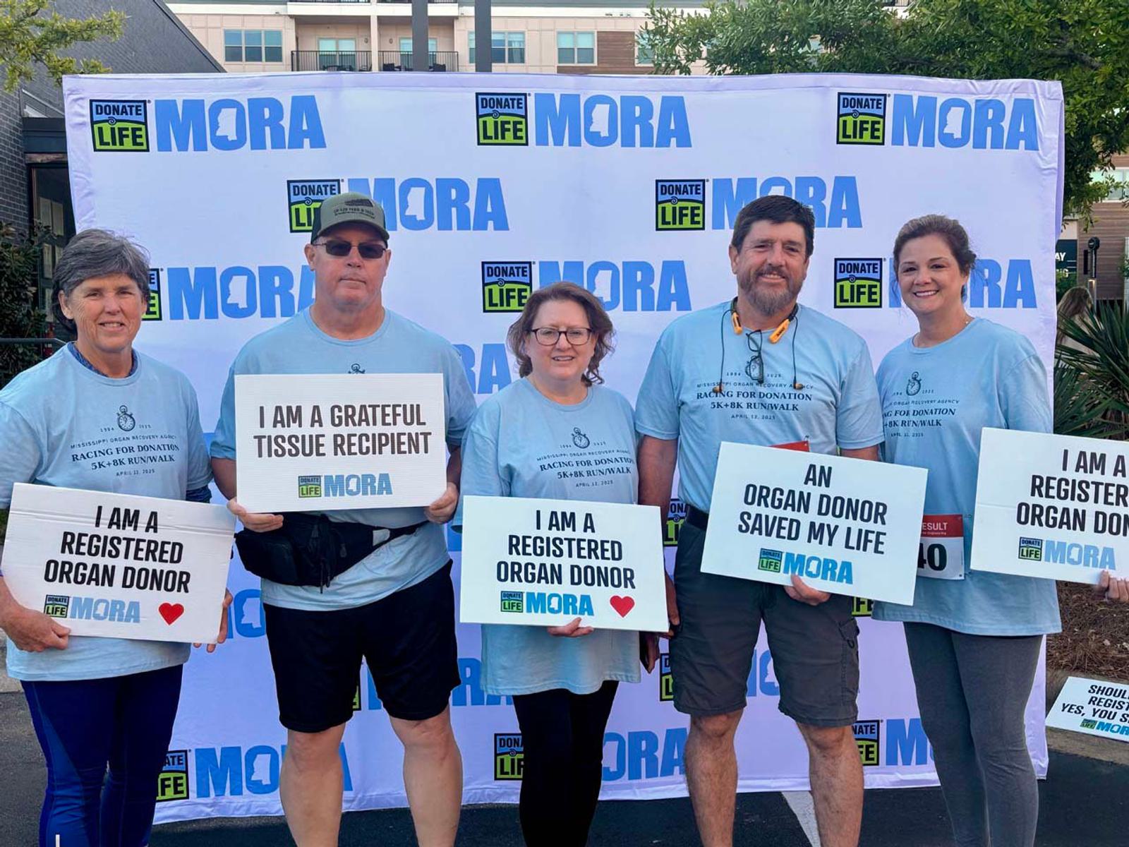 Five people stand in front of a MORA and Donate Life banner, holding signs about organ donation and wearing matching blue event shirts, smiling and showing support for organ and tissue donation.