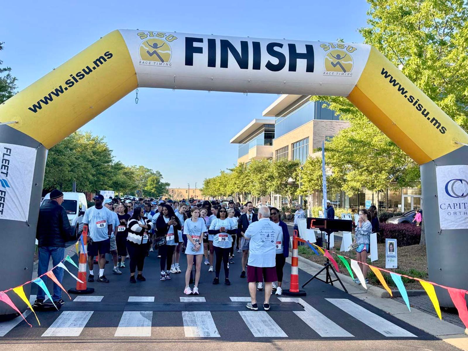 A group of people gather at the starting line under a large yellow “FINISH” archway at an outdoor race event, with colorful flags and trees lining the street.