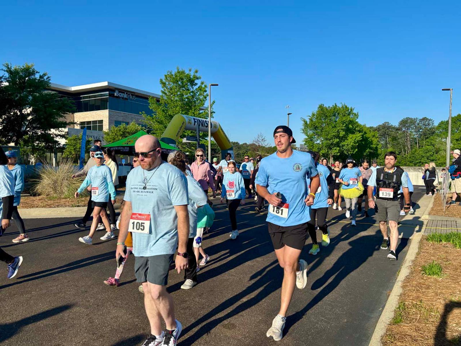 A group of people in numbered bibs and light blue shirts participate in an outdoor charity walk or run on a sunny day. Some are walking, some are jogging, with trees and a building in the background.