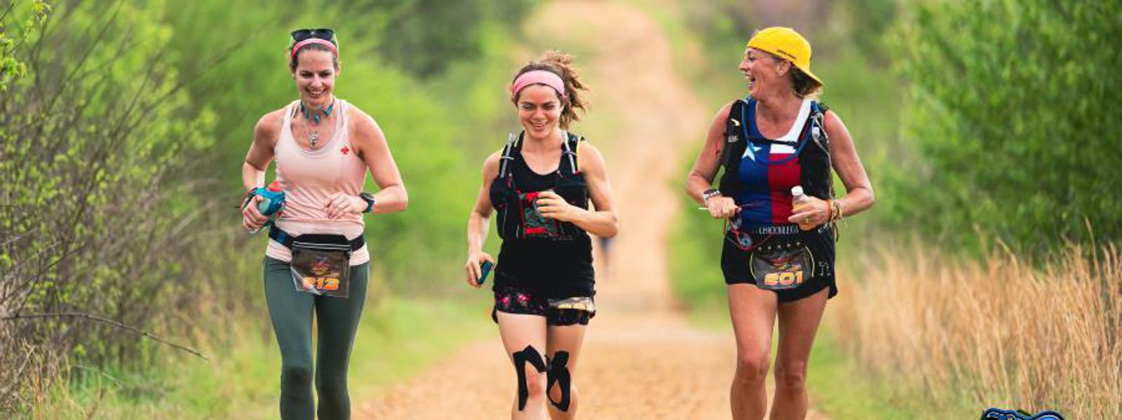 Three women are running together on a dirt trail surrounded by greenery, smiling and wearing athletic gear with race bibs, on a bright day.