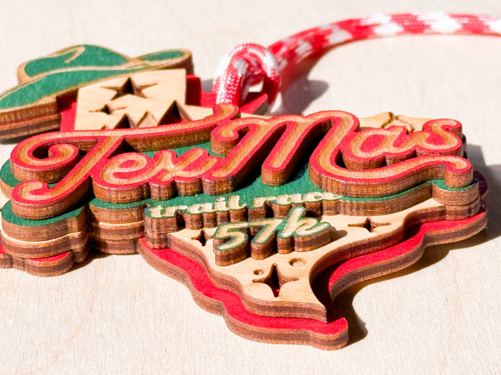 A wooden medal shaped like Texas reads "TexMas trail race 57k" in red, green, and yellow letters, with a red and white checkered ribbon attached.