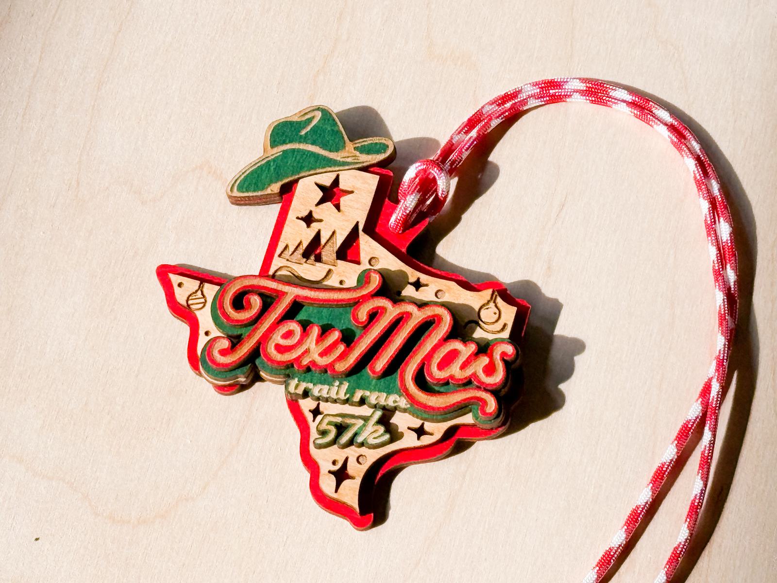A wooden medal shaped like Texas features the words "TexMas trail race 5/7k," decorated with a green cowboy hat, stars, and a red and white rope lanyard.