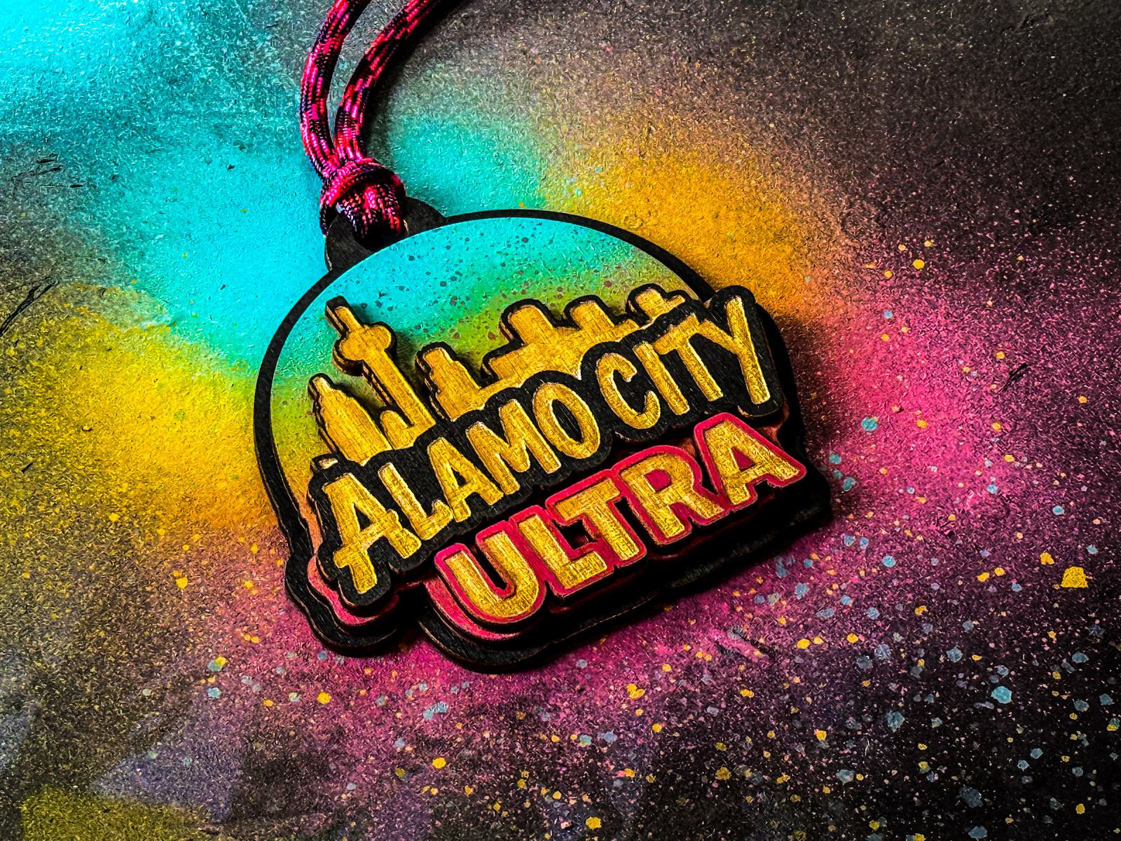 A colorful medal with the words "Alamo City Ultra" and a city skyline, set against a vibrant background of splattered yellow, blue, pink, and black paint.