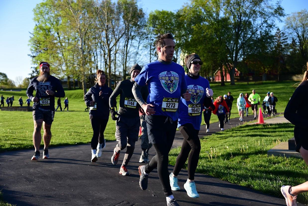 A group of runners wearing race bibs and athletic gear jogs along a park path on a sunny day, with green grass, trees, and other participants visible in the background.