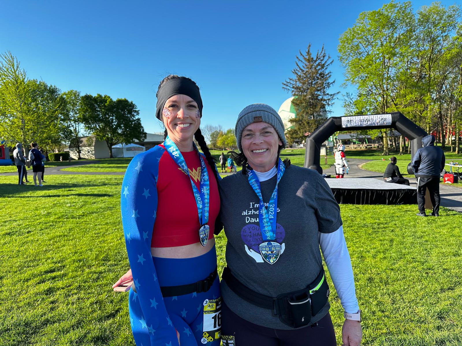 Two smiling women stand outside on grass after a race, each wearing a finisher’s medal. One is dressed in a superhero costume; the other wears a T-shirt and beanie. Trees and an inflatable race arch are visible in the background.