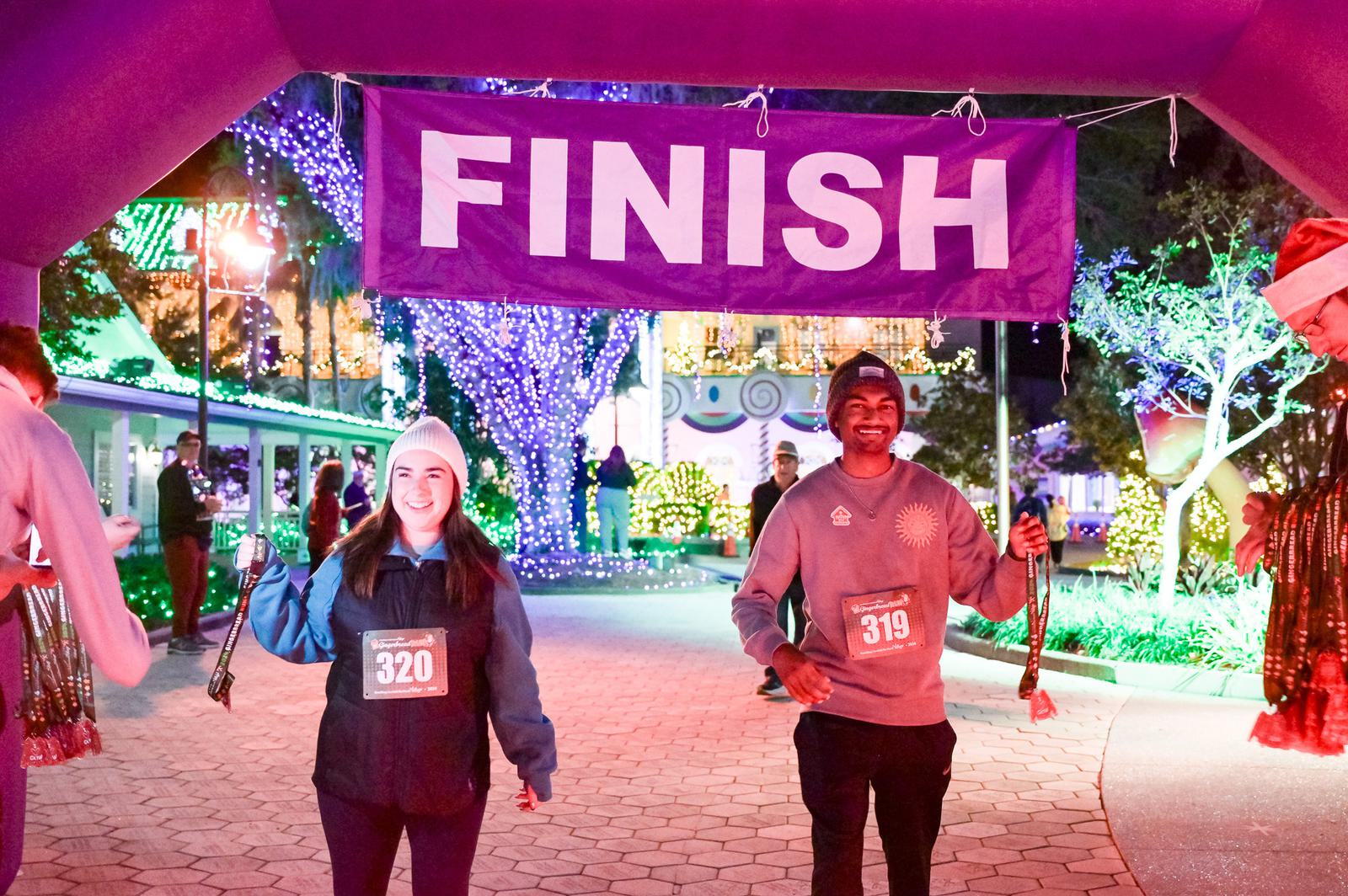 Two runners, wearing race bibs 320 and 319, smile as they cross a finish line under purple lights at night, each holding medals. Colorful holiday lights and decorations fill the background.