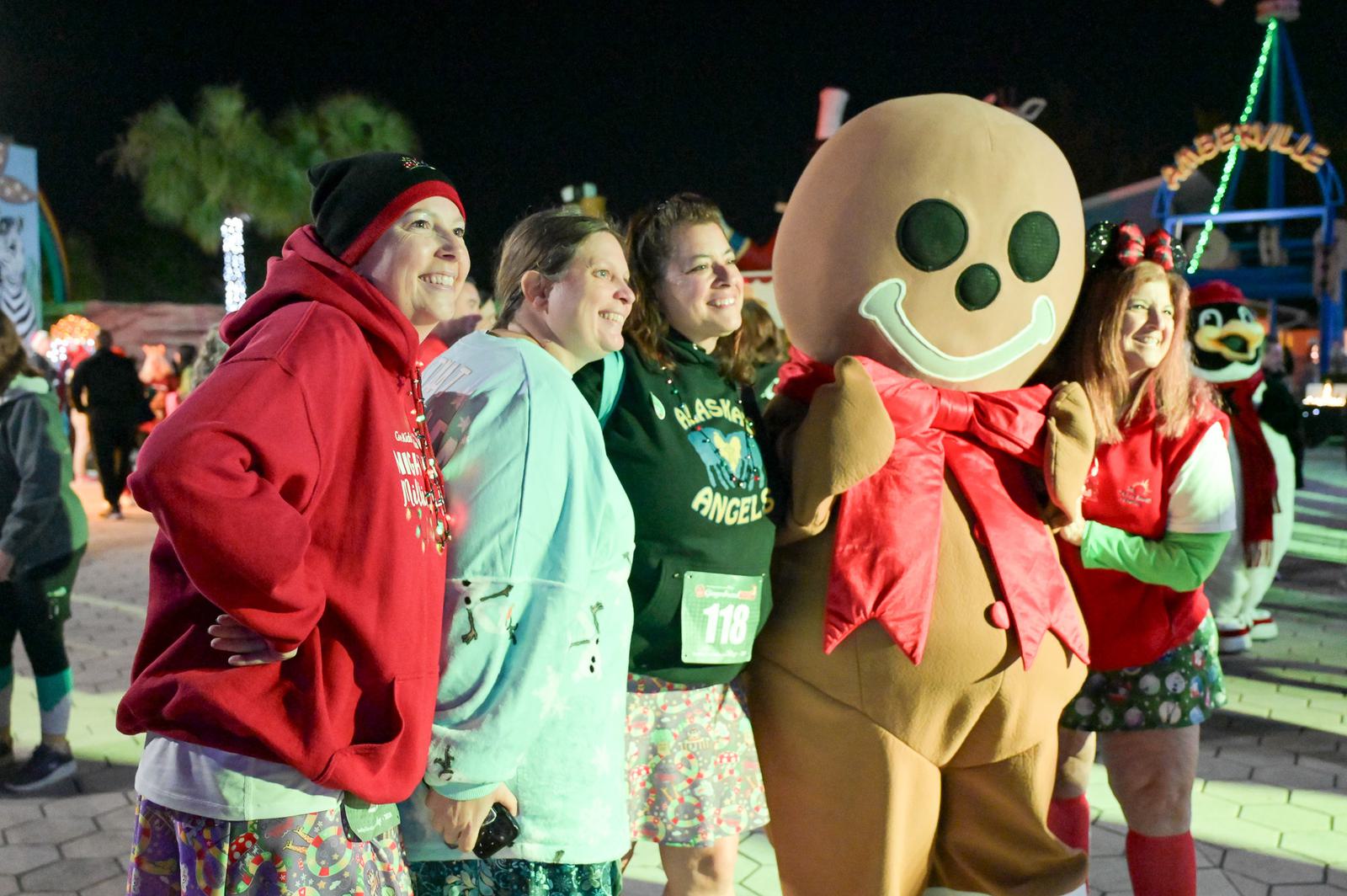 A group of four women pose and smile next to a person in a large gingerbread man costume, all dressed festively, at an outdoor nighttime event with holiday decorations.
